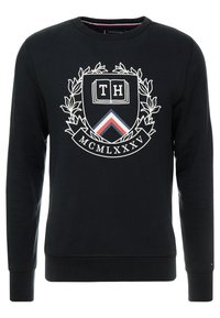 Sort sweatshirt med rund halsudskæring, der har et centralt skjoldlogo med "TH", laurbærdetalje og et trekantet mønster i rød og blå.