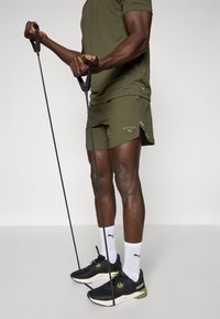 Ensemble de sport olive avec un short et un t-shirt, arborant un logo. Comprend des baskets noires, des chaussettes blanches et des bandes de résistance pour l'exercice.