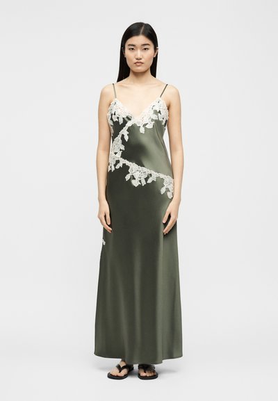 Γυναίκα που φορά ένα μακρύ σατέν φόρεμα σε ελιά πράσινο με λευκή δαντελένια διακόσμηση, λεπτές τιράντες στους ώμους και μαύρα επίπεδα σανδάλια, στέκεται μπροστά σε απλό φόντο.