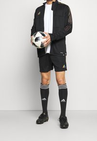 Zwarte sportjack met regenbooglijnen, witte T-shirt, zwarte shorts, zwart-wit voetbal, zwarte voetbalschoenen en zwarte sokken met strepen.