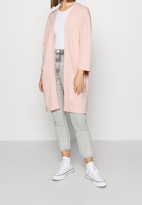 Cárdigan de color rosa claro, de apertura frontal, con mangas tres cuartos, llevado sobre una blusa blanca y combinado con jeans gris claro y zapatillas blancas.
