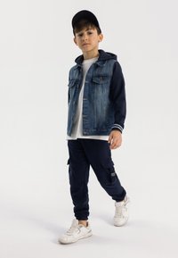 Denimjack met blauwe mouwen, wit T-shirt, marineblauwe joggers met zakken, en witte sneakers met een zwarte pet. Schoon, modern esthetisch.