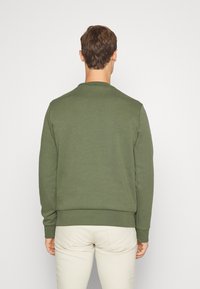 Muž oblečený v olivově zelené mikině s dlouhým rukávem a světle béžových kalhotách, stojící zády k útěrnému světlému pozadí.