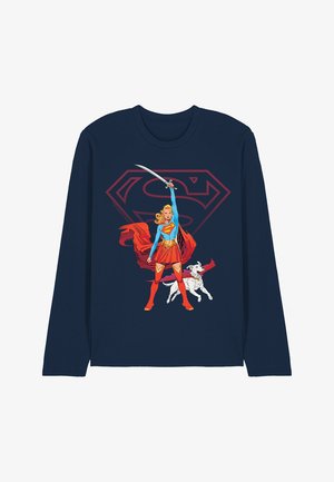 Supergirl in blue and red costume holding a sword aloft, standing next to a white dog, with a large red Superman logo behind her on navy shirt.