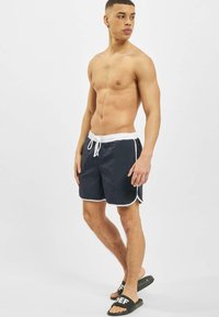 Marine svømmetruser med hvit kant og elastisk midjebånd, med et merkelappdetalj. Båret med svarte sandaler, står modellen uten skjorte.