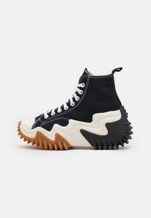 Sort høj sneaker med hvide snørebånd, tyk bølget creme midtersål og takkede brune for- og sorte bagsåler.
