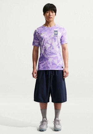 Νέος άνδρας που στέκεται φορώντας μοβ φανέλα ποδοσφαίρου της Κορέας με λουλουδάτο σχέδιο, μαύρο φαρδύ σορτς, γκρι κάλτσες και γκρι αθλητικά παπούτσια σε φωτεινό φόντο.