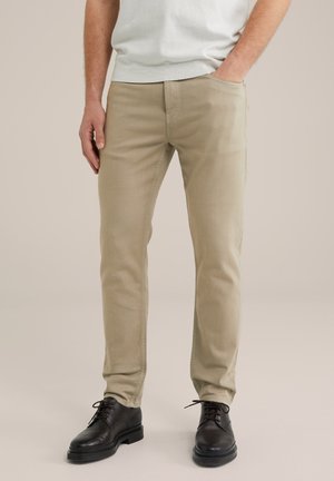 Mand iført beige slim-fit bukser med hånden i lommen og sorte lædersko, stående mod en neutral baggrund.