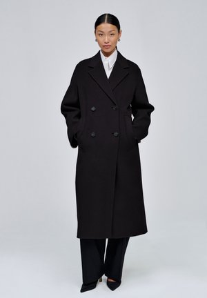 Une femme porte un long manteau noir croisé sur une chemise blanche et un pantalon noir, devant un fond de studio clair.