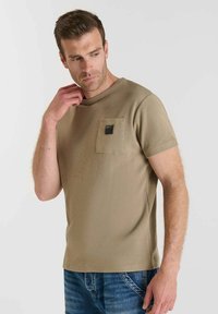 Camiseta texturizada marrón con cuello redondo y mangas cortas. Presenta un bolsillo en el pecho con una pequeña etiqueta negra. Combinada con jeans azules.