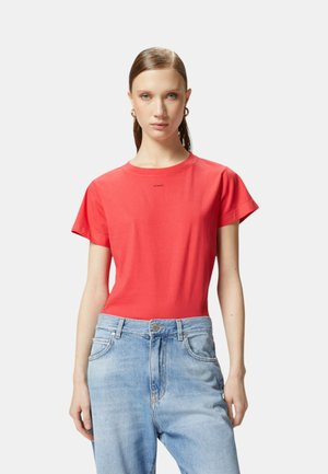 Maglietta rossa in cotone con maniche corte e colletto a girocollo, caratterizzata da un piccolo logo stampato vicino al colletto. Indossata con jeans azzurri chiari.