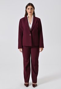 Frau steht in einem bordeauxroten Anzug mit einem Ein-Knopf-Blazer, einer weißen Bluse und bordeauxroten spitzen Schuhen vor einem schlichten hellen Hintergrund.