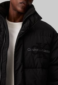 Fekete, steppelt dzseki, elöl cipzárral, magas nyakkal, és hímzett "Calvin Klein Jeans" logóval a mellkasán. Puha, steppelt textúrájú.