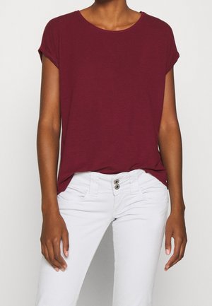 Personne portant un haut bordeaux à manches courtes et un pantalon blanc, debout les bras détendus le long du corps devant un fond clair uni.