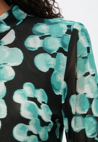 Blusa preta com um padrão abstrato em turquesa com formas redondas, feita de um tecido leve, translúcido e com uma leve textura.