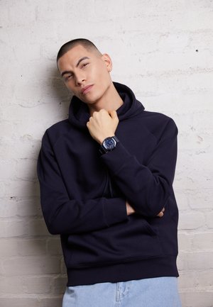 Granatowy hoodie o teksturowanym wykończeniu, z kieszenią kangurką i ściągaczami przy rękawach. W komplecie z ciemnym zegarkiem i jasnoniebieskimi dżinsami.