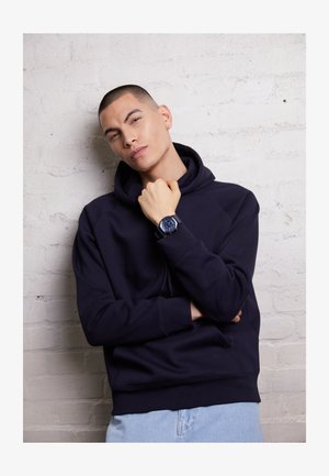 Navy hoodie with a textured finish, featuring a kangaroo pocket and ribbed cuffs. Paired with a dark watch and light blue jeans.