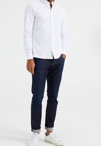 Biała, długorękawowa koszula zapinana na guziki noszona z ciemnymi jeansami, o dopasowanym kroju, z kontrastującymi przeszyciami i podwiniętymi mankietami, w zestawie z białymi trampkami.