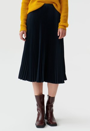 Femme portant un pull jaune moutarde, une jupe midi plissée noire et des bottines à talons marron foncé sur un fond uni.