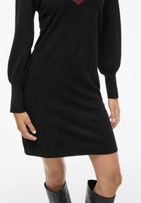 Czarna dzianinowa sukienka z długimi, bufkami i głębokim bordowym wzorem w kształcie litery V na piersi. Kończy się nad kolanem. Gładka faktura.