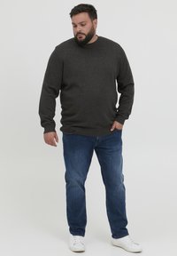 Pull gris foncé à col rond et manches longues, associé à un jean en denim bleu et des baskets blanches sur fond uni.