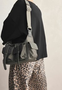 Zielona nylonowa torba na ramię z wieloma przednimi kieszeniami, regulowanym paskiem i detalami przeszycia. Noszona z spodniami w zwierzęcy wzór i czarnym topem.