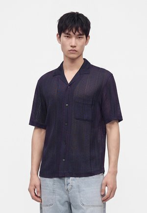 Jeune homme portant une chemise sombre à motifs à manches courtes et un jean bleu clair, debout devant un fond blanc uni.
