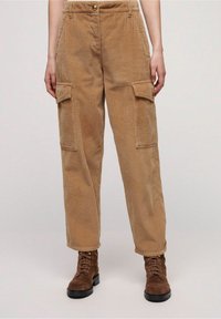 Koeienkleurige corduroy cargobroek met een getextureerd oppervlak, voorzien van twee zijzakken en een knoopsluiting. Gecombineerd met bruine veterschoenen.