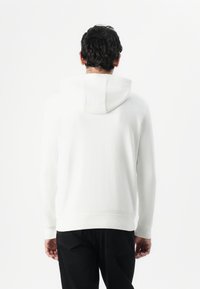 Weißer Hoodie mit Kordelzugkapuze, langen Ärmeln, gerippten Bündchen und Saum. Getragen mit schwarzen Jeans, von hinten betrachtet, vor einem schlichten Hintergrund.