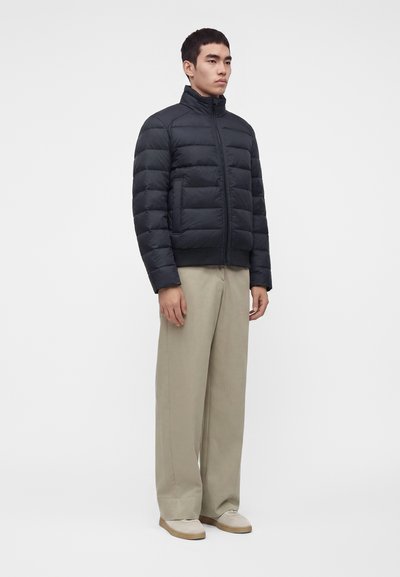 Homme portant une doudoune noire, un pantalon beige à jambes larges et des chaussures de couleur claire, debout sur un fond blanc uni.