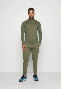 Mężczyzna stojący z rękami w kieszeniach, ubrany w oliwkowozielony długi rękaw z zamkiem oraz bojówki typu jogger, niebiesko-czarne sneakersy.