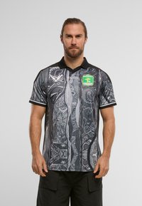 Fekete és szürke mintázatú pólóing gallérral, absztrakt mintázatokkal és zöld emblémával a mellkasán. Rövid ujjak és testhez simuló fazon.