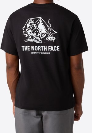 Person iført sort T-shirt med hvid campinggrafik af person ved bål og telt, og teksten "THE NORTH FACE NEVER STOP EXPLORING" på ryggen.