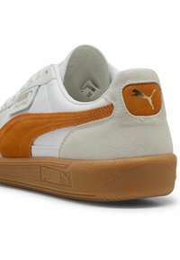 Hvide sneakers med orange ruskindsdetaljer og en tekstureret gummisål. Viser et guld Puma-logo på hælen og er lavet af læder.