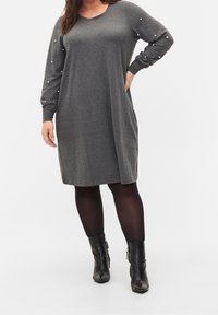 Graues, langärmeliges Kleid mit lockerem Schnitt, das Perlenakzente an den Ärmeln aufweist. Getragen mit schwarzen Strumpfhosen und Ankle Boots. Glatte Textur.