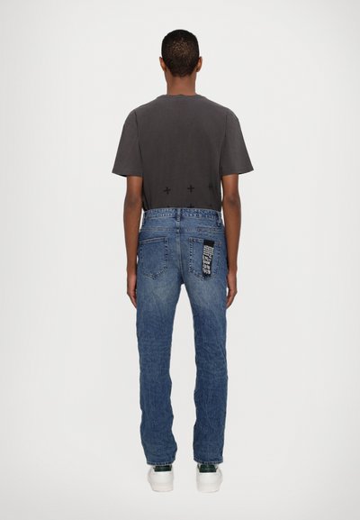 Graues T-Shirt mit kurzen Ärmeln und Rückenmotiv, kombiniert mit blauen Jeans aus Denim, die ein schwarzes Patch an der Gesäßtasche und einen lockeren Schnitt haben.