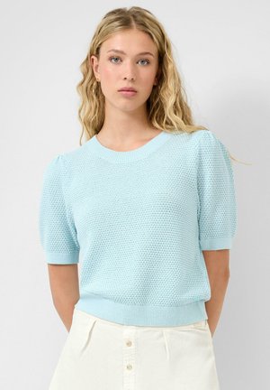 Junge Frau mit langem blondem Haar trägt einen hellblauen, strukturierten Kurzarm-Pullover und weiße, hoch taillierte Hose mit Knöpfen.