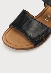Black leather sandal with two wide straps, showcasing a smooth texture and a cushioned tan footbed. Lightweight sole with rounded shape.