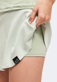 Main tenant le côté d'une jupe de sport vert clair texturée avec des shorts intégrés et une étiquette de marque noire Endless.