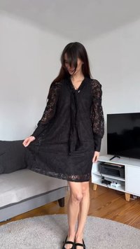 Juoda nėriniuota suknelė su ilgomis rankovėmis ir kaklo raišteliu. Suknelė yra laisvo silueto, su nėriniuotomis bangelėmis apačioje. Dėvima kartu su juodais butais batais.