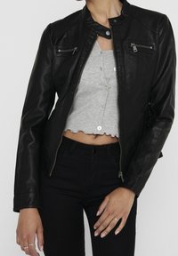 Black leather biker jacket with a fitted design, high collar, and zippered pockets. Worn over a gray cropped shirt and black jeans.
