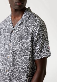 Short-sleeve shirt in black and white with a swirling text pattern featuring "GUESS." Includes a chest pocket and wooden buttons.
