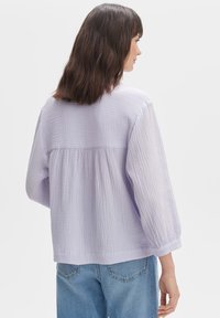 Blusa oversize in lavanda chiaro con tessuto texturizzato, schiena arricciata e maniche a palloncino, abbinata a jeans azzurri.