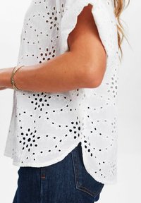 Blusa bianca ricamata con dettagli a occhio di pernice, maniche corte e orlo scallop. Abbinata a un tessuto denim blu scuro.