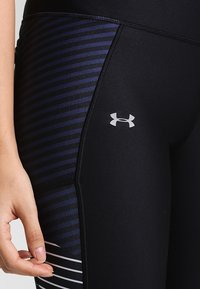 Černé legíny s přiléhavým střihem, které mají na boku námořnickou modrou pruhovanou vzor a stříbrné logo Under Armour blízko kyčle.