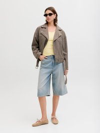 Vrouw met schildpadprint zonnebril, taupekleurig suède jasje, geel topje, knielange spijkershorts en beige ballerina's.