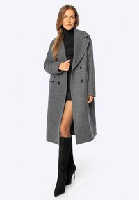 Cappotto di lana grigio con una vestibilità lunga e ampia, grandi rever, design doppiopetto e tasche laterali. Indossato sopra un top nero e una gonna corta.