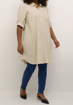 Femme portant une robe chemise longue beige avec manches retroussées, un jean skinny bleu et des talons slingback noirs à bout pointu, debout contre un mur uni.