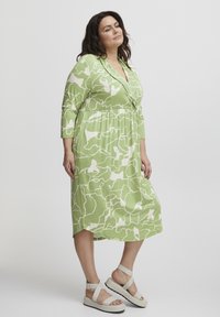Vestido midi verde con un patrón abstracto blanco, mangas largas, escote en V y cintura elástica. Combina con sandalias de plataforma de color claro.