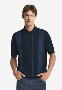 Navyrandig polo i mjukt tyg, med korta ärmar, knapplist och varierande blå nyanser i vertikala linjer.